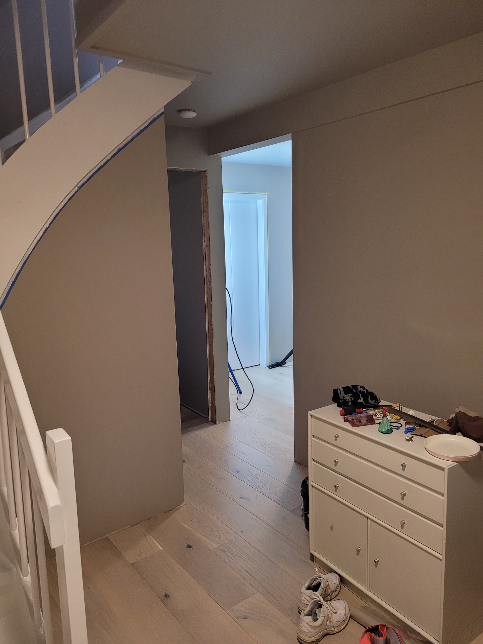 Korytarz w trakcie odświeżania: ściany pomalowane na beżowo, widoczne schody z białą balustradą zabezpieczone taśmą malarską, komoda z narzędziami i akcesoriami malarskimi, podłoga drewniana.