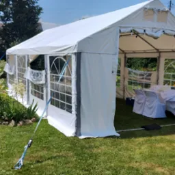 Biały namiot imprezowy rozstawiony na trawniku, widoczne stoły i krzesła wewnątrz, z boku huśtawka ogrodowa i kwitnące rośliny.