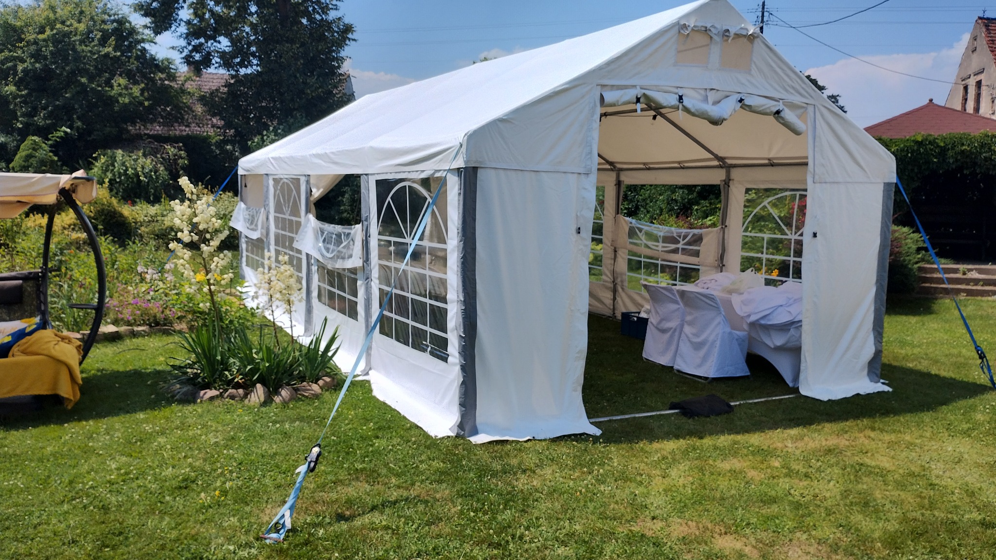 Biały namiot imprezowy rozstawiony na trawniku, widoczne stoły i krzesła wewnątrz, z boku huśtawka ogrodowa i kwitnące rośliny.