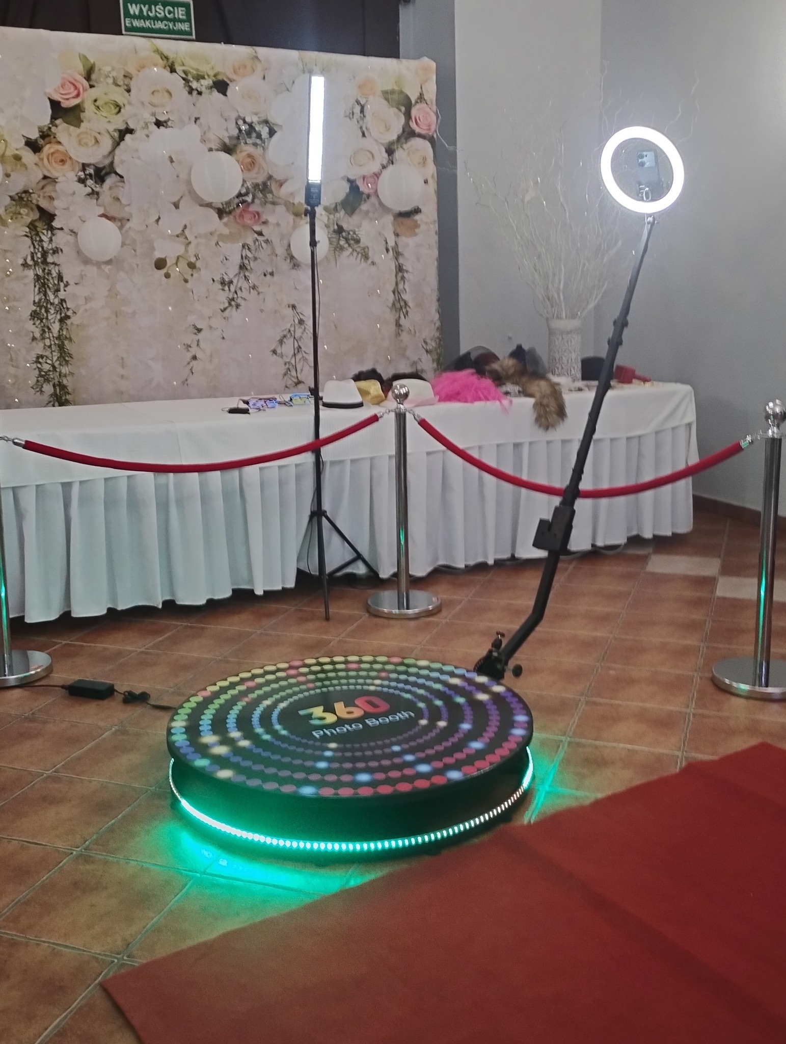 Okrągła platforma 360 Photo Booth z kolorowymi światłami LED na podłodze z płytek, w tle dekoracja kwiatowa i rekwizyty na stole, oświetlenie studyjne.