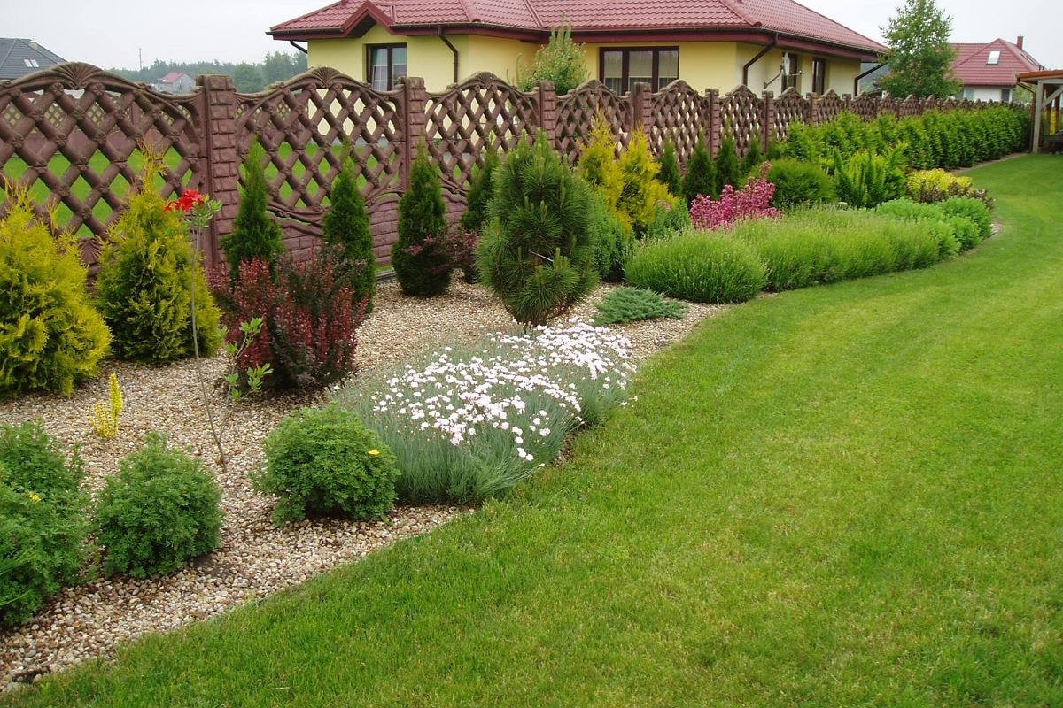 Ukształtowany ogród przydomowy z dekoracyjnym, ażurowym ogrodzeniem, ozdobnymi krzewami i kwiatami na żwirowym podłożu oraz równo przystrzyżonym trawnikiem.