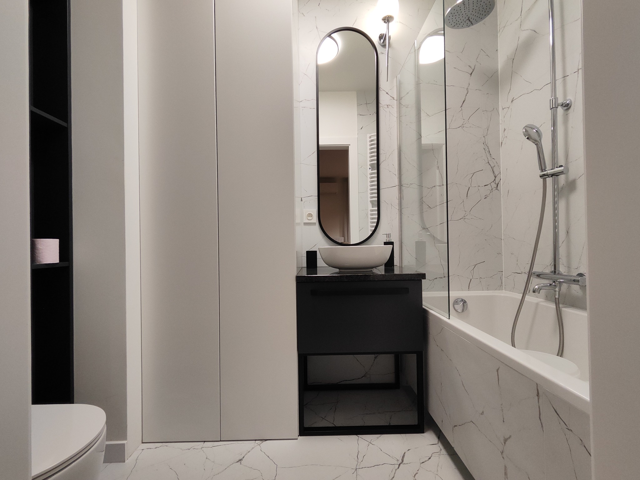 Nowoczesna łazienka z marmurowymi płytkami, owalnym lustrem i czarną szafką pod umywalką, widoczna bateria prysznicowa z deszczownicą i wanna.