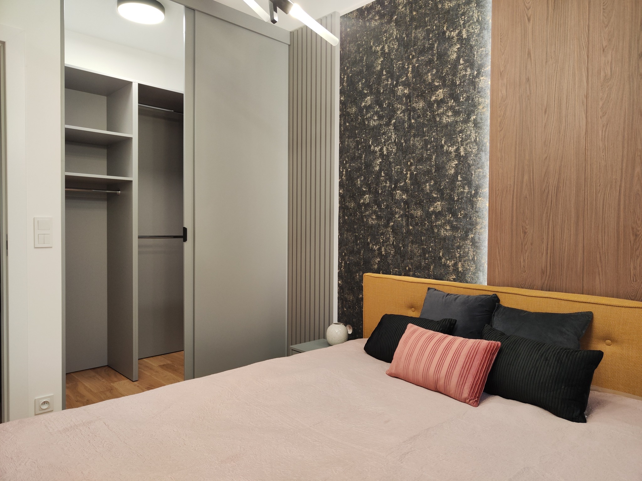 Sypialnia z łóżkiem z żółtym zagłówkiem i dekoracyjnymi poduszkami, na ścianie tapeta w odcieniach szarości i złota, obok przesuwne drzwi do garderoby.