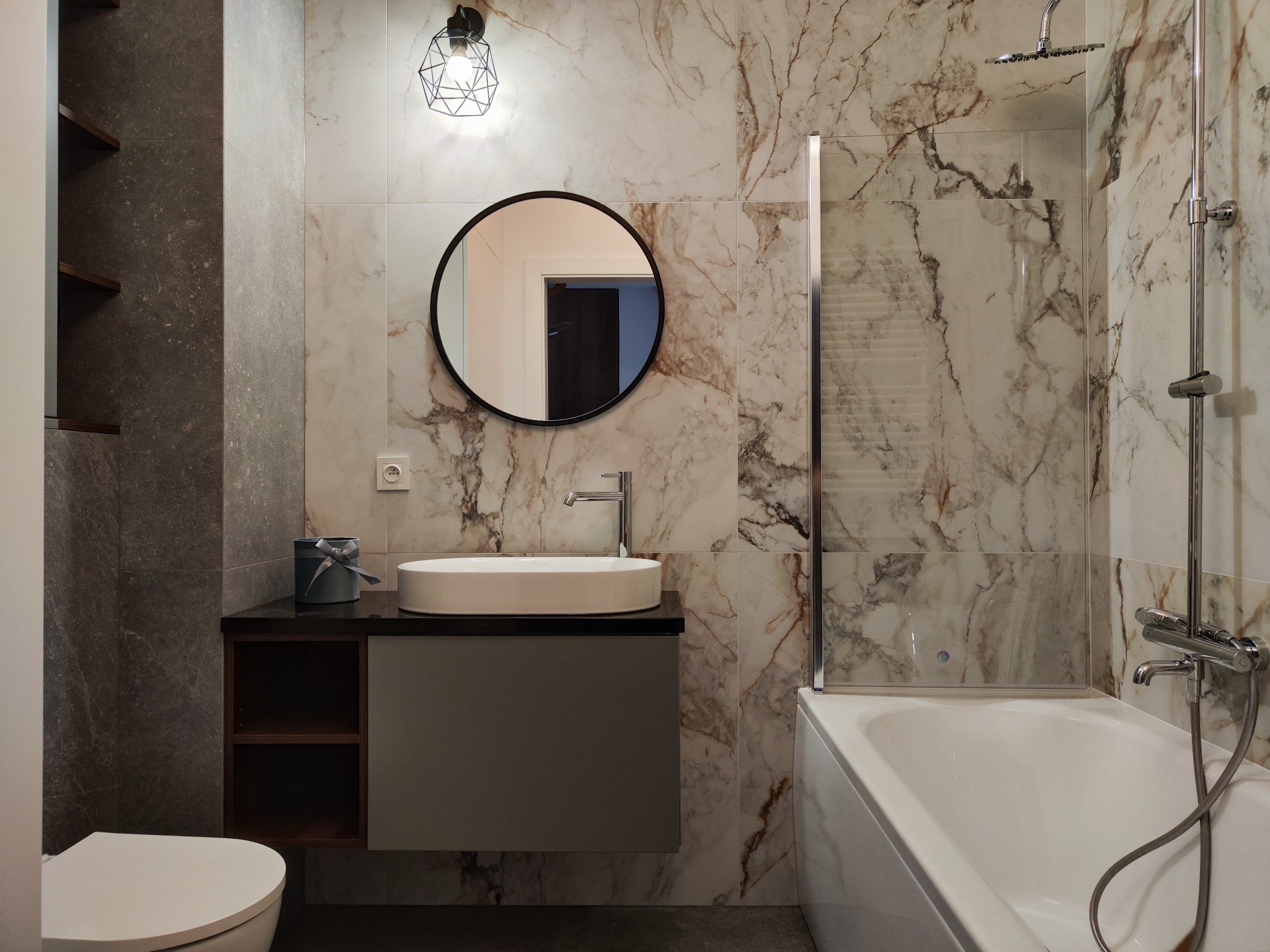 Nowoczesna łazienka z marmurowymi płytkami, okrągłym lustrem, owalną umywalką nablatową i geometryczną lampą sufitową.