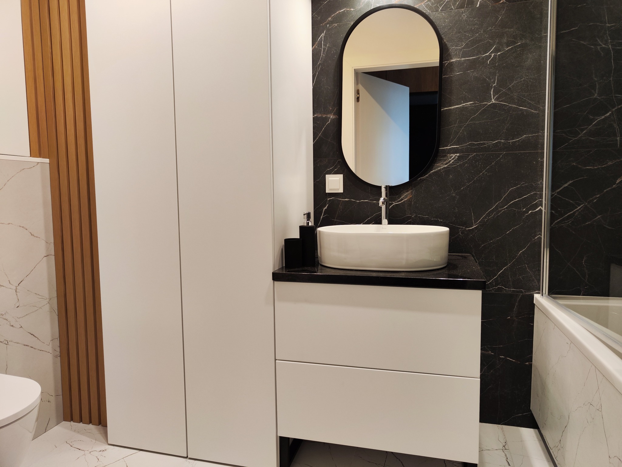 Nowoczesna łazienka z owalnym lustrem, białą umywalką nablatową i czarnym blatem na szafce z szufladami, ściany wyłożone czarnym marmurem z białymi żyłami, widoczna część wanny.