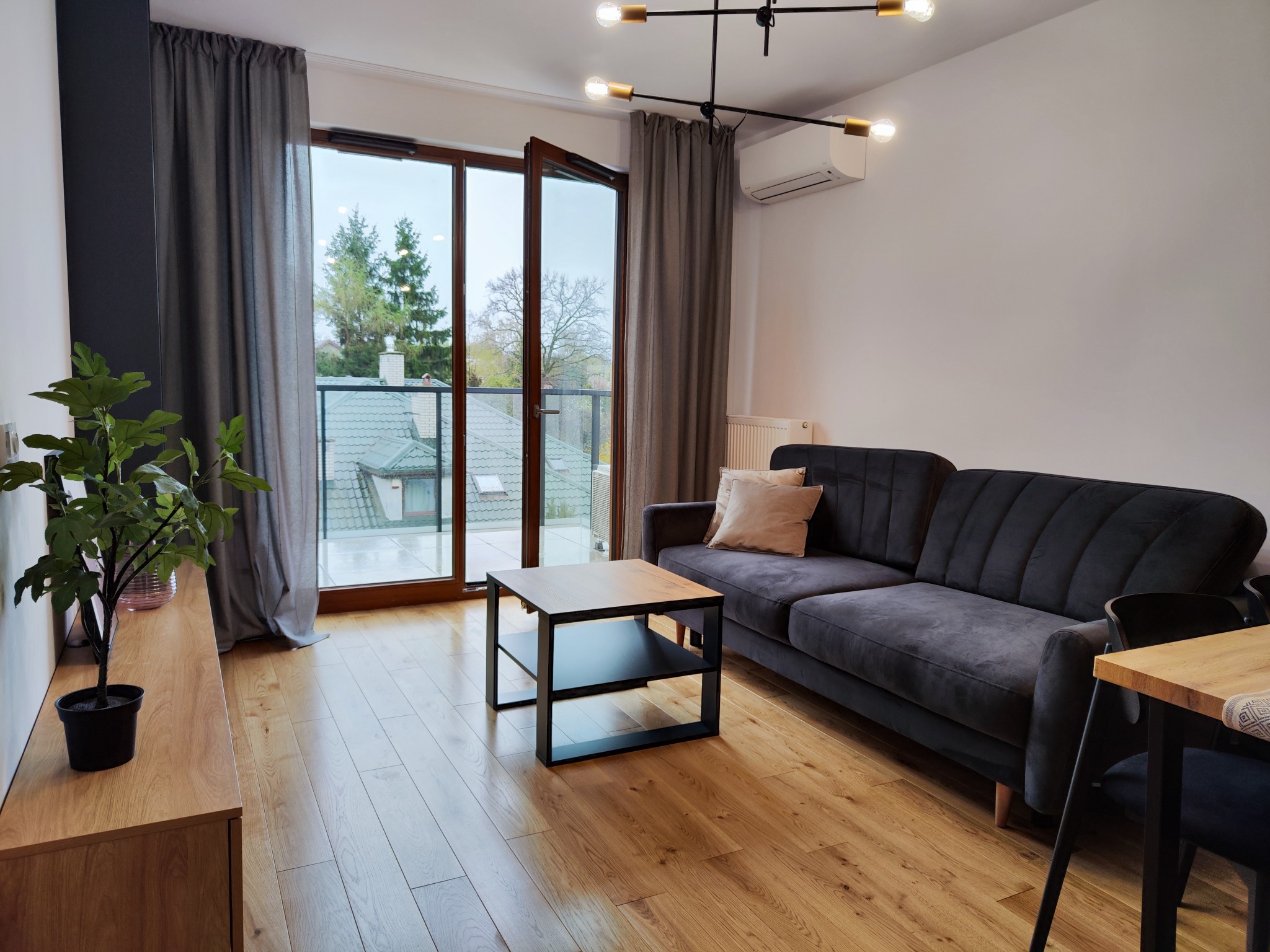 Jasne wnętrze salonu z drewnianą podłogą, szarą sofą, stolikiem kawowym i widokiem na balkon przez otwarte szklane drzwi.