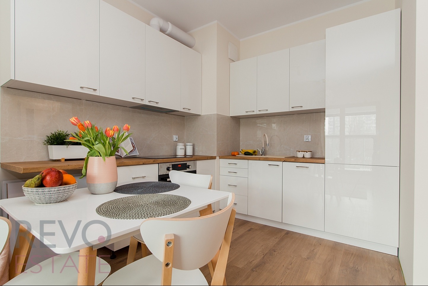 Nowoczesna, jasna kuchnia z białymi, lakierowanymi szafkami, drewnianym blatem, marmurową ścianą i stołem z owocami i tulipanami w wazonie.