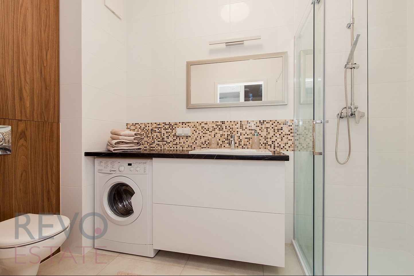 Funkcjonalna łazienka w Warszawie z pralką obok umywalki, wykończona mozaiką i białymi płytkami, kabina prysznicowa ze szklanymi drzwiami.