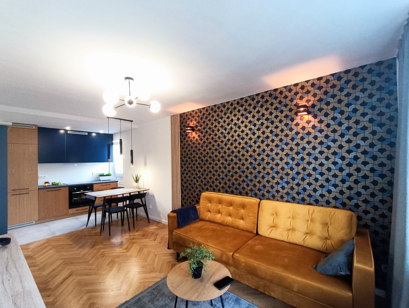 Salon z otwartą kuchnią, parkiet w jodełkę, musztardowa sofa, ściana z geometrycznym wzorem, nowoczesne oświetlenie.