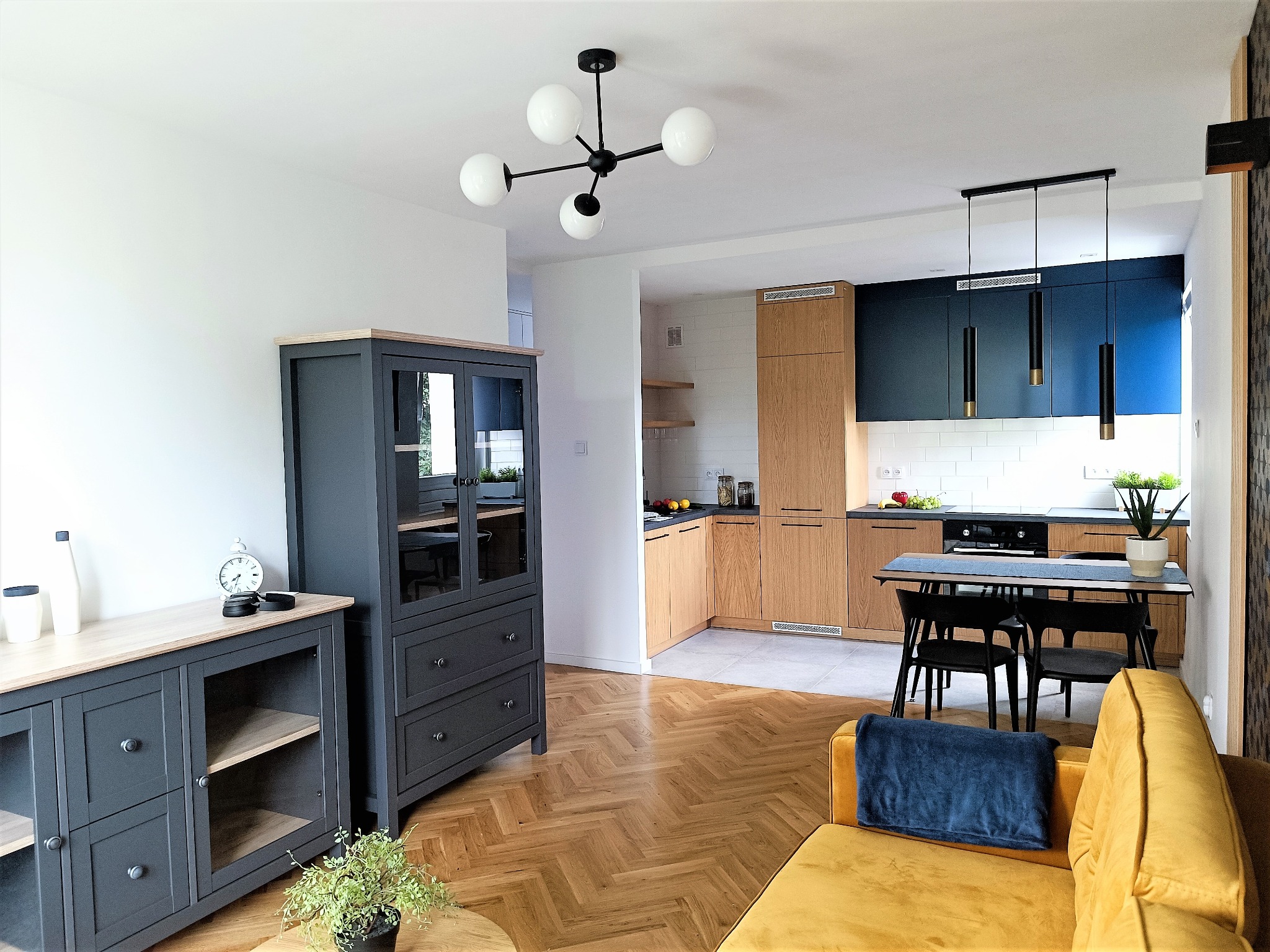 Jasne wnętrze mieszkania z parkietem w jodełkę, szarymi meblami, żółtą sofą i otwartą kuchnią z drewnianymi i granatowymi szafkami oraz białymi płytkami na ścianie.