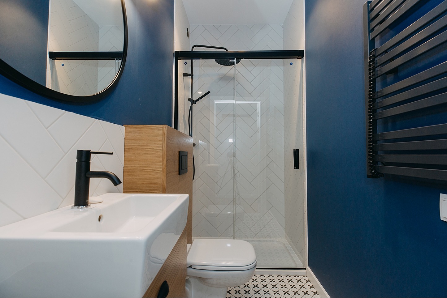 Nowoczesna łazienka z granatowymi ścianami, białą umywalką z czarną armaturą, kabiną prysznicową z białymi płytkami ułożonymi w jodełkę oraz czarnym grzejnikiem drabinkowym.