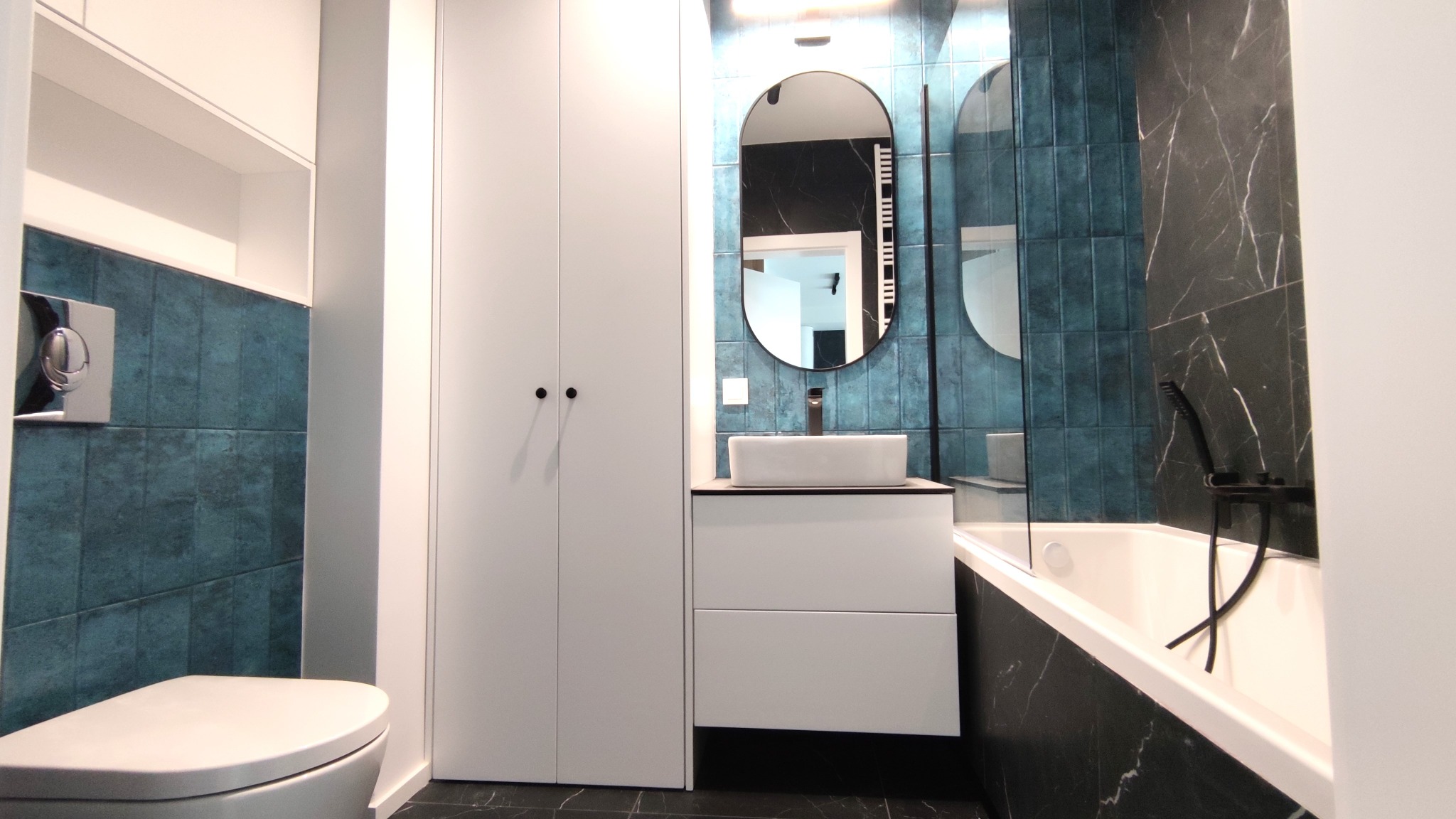 Nowoczesna łazienka z turkusowymi płytkami i czarnym marmurem, owalne lustro nad prostokątną umywalką, biała szafka i zabudowa, czarna armatura.