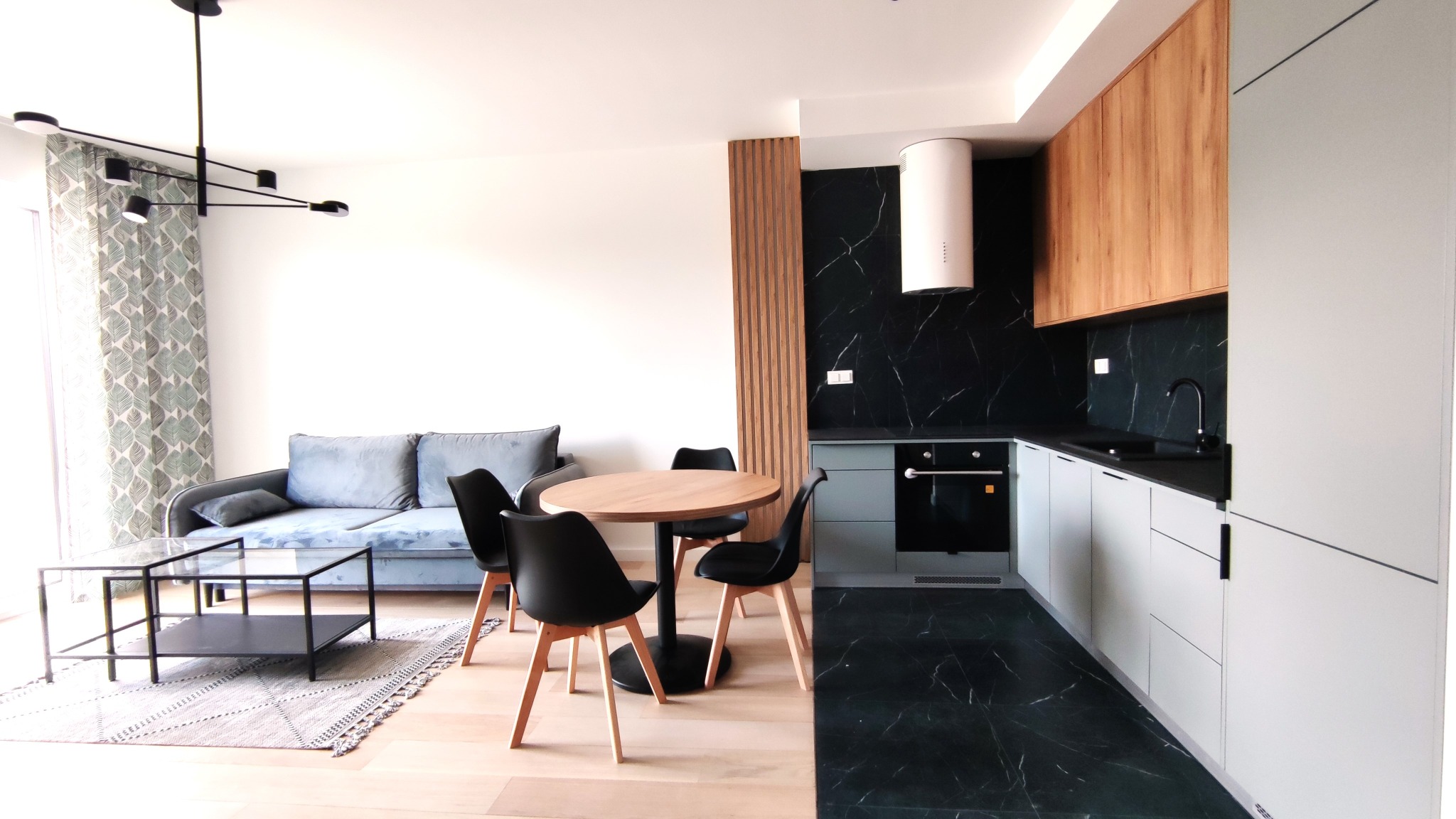 Nowoczesne wnętrze mieszkania z aneksem kuchennym, drewnianym stołem i czarnymi krzesłami, szarą sofą i stolikiem kawowym na wzorzystym dywanie.