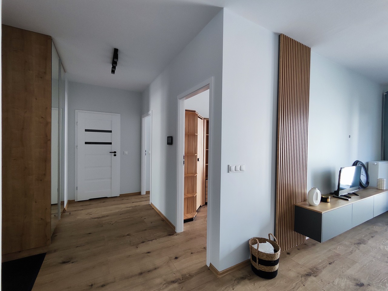 Nowoczesny przedpokój z drewnianą podłogą, pionowymi panelami ściennymi z drewna, białą ścianą i minimalistyczną szafką RTV w kolorze szarym.