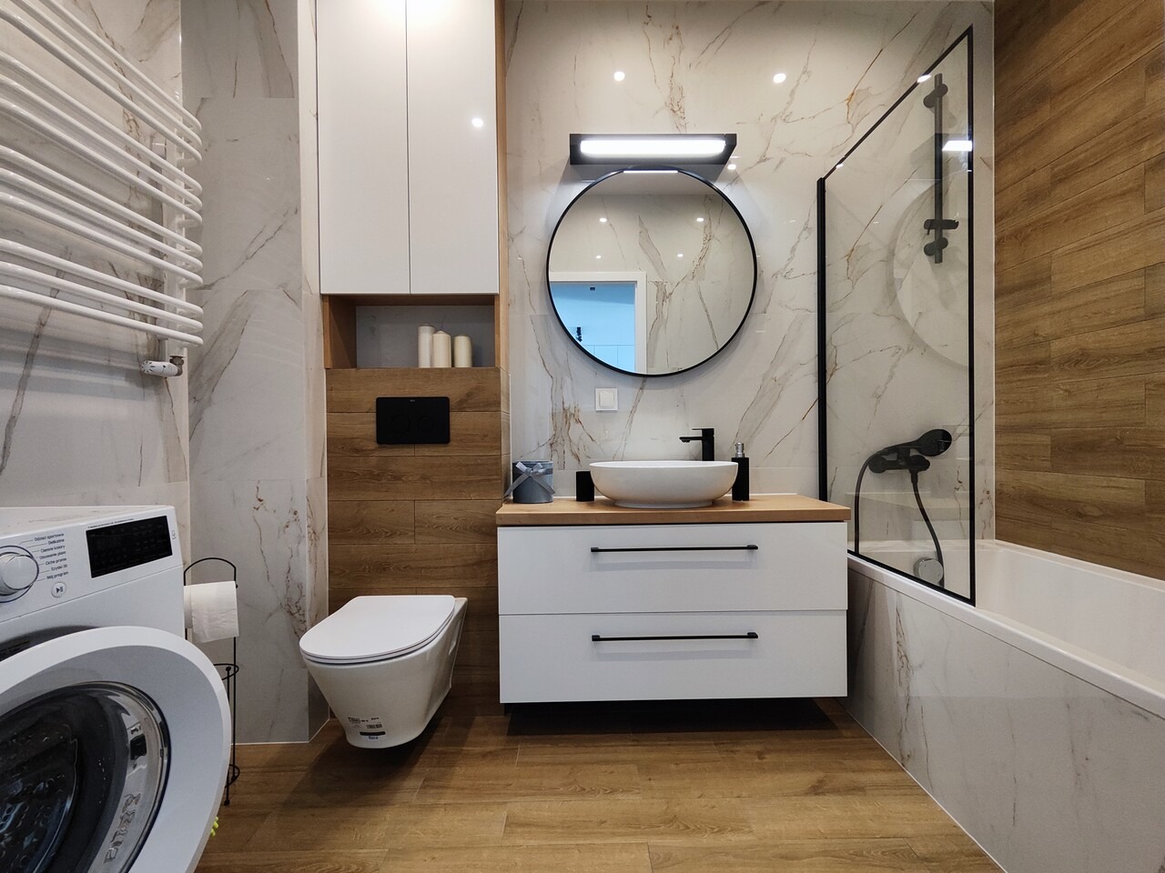 Nowoczesna łazienka z marmurowymi płytkami, okrągłym lustrem z czarną ramą, białą umywalką nablatową, baterią w kolorze czarnym, drewnopodobną podłogą, pralką oraz grzejnikiem drabinkowym.