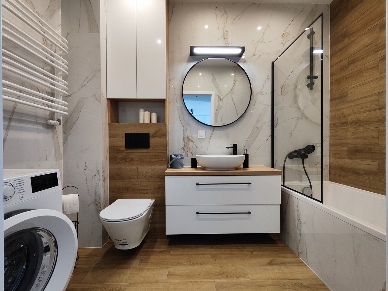 Nowoczesna łazienka z marmurowymi płytkami, okrągłym lustrem z podświetleniem LED, białą umywalką nablatową, wiszącą toaletą i pralką z suszarką