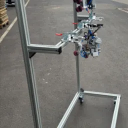 Stojak z profili aluminiowych na kółkach z zamontowanym, skomplikowanym oprzyrządowaniem pneumatycznym z przyssawkami i zaciskami, na zewnątrz.