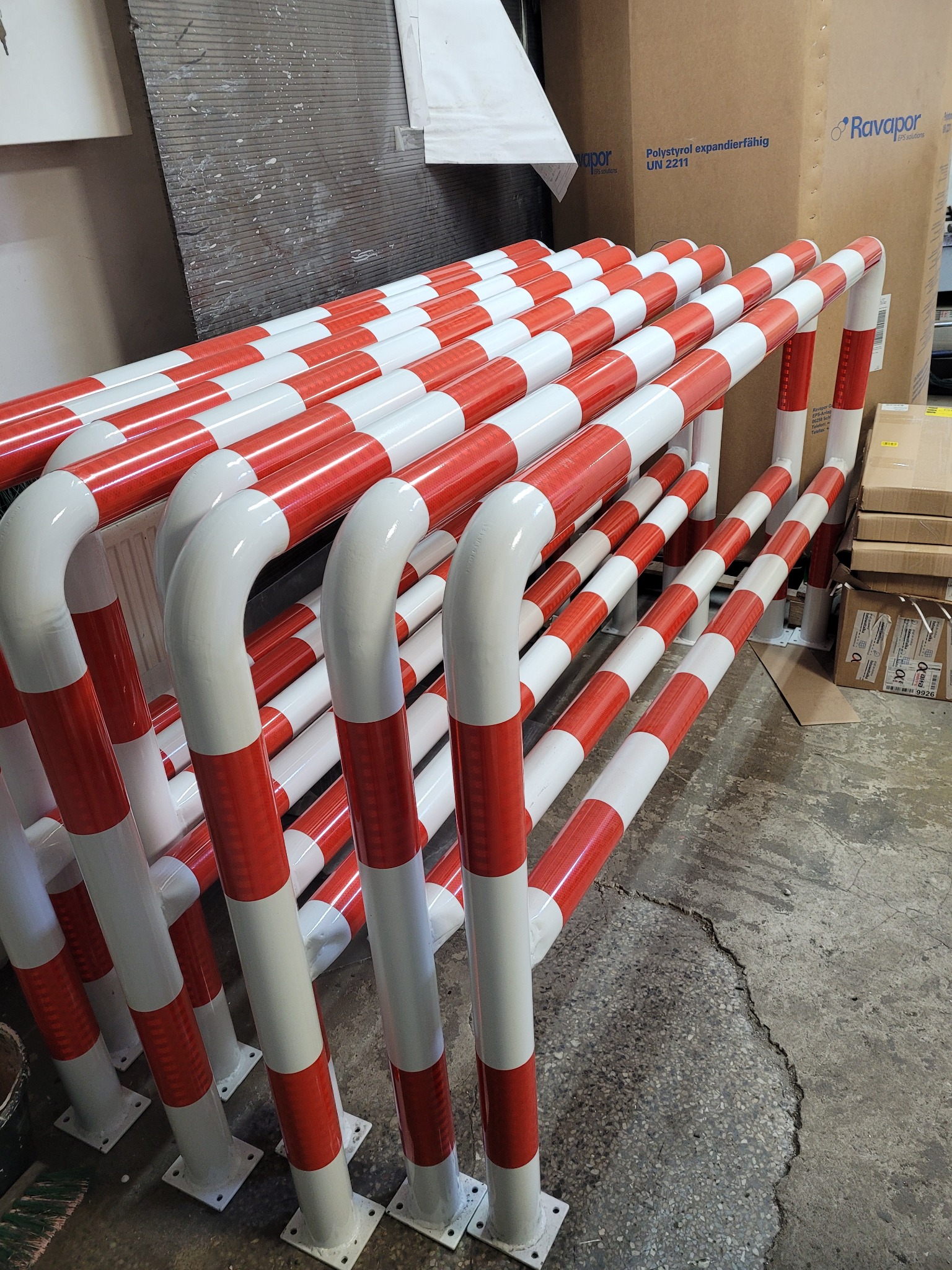Stojaki ochronne z rur stalowych pomalowanych w biało-czerwone pasy, ułożone w magazynie na betonowej podłodze, widoczne kartony w tle.