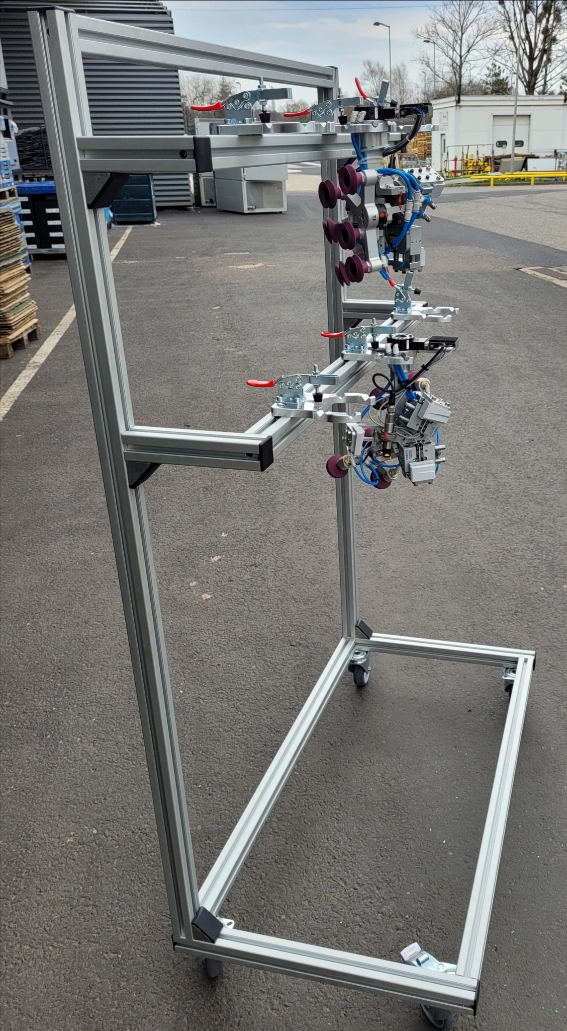 Stojak z profili aluminiowych na kółkach z zamontowanym, skomplikowanym oprzyrządowaniem pneumatycznym z przyssawkami i zaciskami, na zewnątrz.