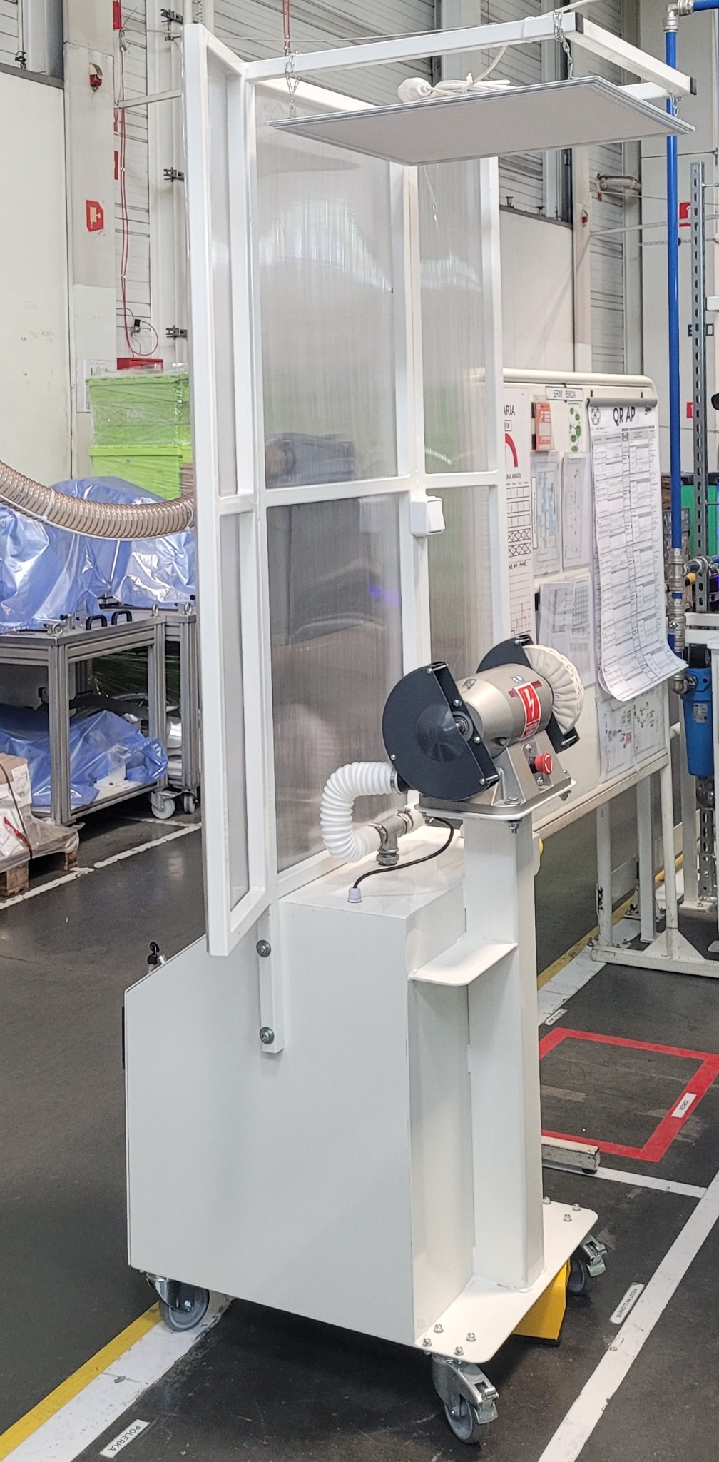 Mobilna stacja robocza z szlifierką polerską, wyposażona w przezroczystą osłonę i oświetlenie, umieszczona w hali produkcyjnej.