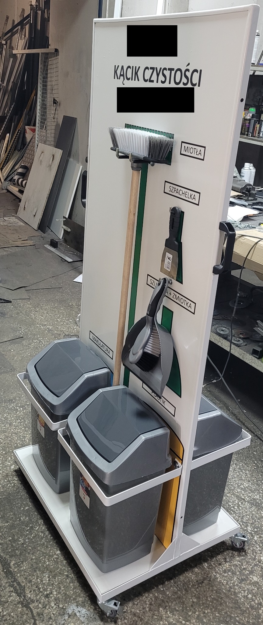 Mobilny stojak na narzędzia i pojemniki do segregacji śmieci z napisem 'Kącik Czystości', wyposażony w miotłę, szpachelkę, szufelkę ze zmiotką oraz pojemniki na makulaturę i plastik, umieszczony...