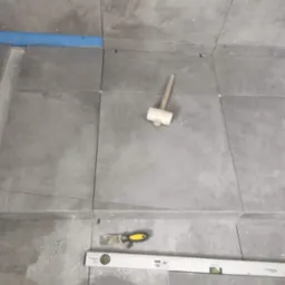 Szare płytki podłogowe w trakcie układania w łazience, widoczny odpływ liniowy, poziomica i gumowy młotek.