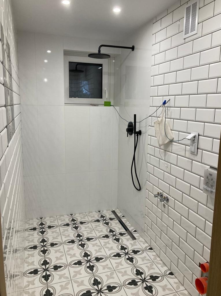 Wykończona kabina prysznicowa z czarną armaturą, białą glazurą typu metro, wzorzystą podłogą w odcieniach szarości i czerni, odpływ liniowy, okno z widokiem na zewnątrz, widoczny woreczek na klamerce.