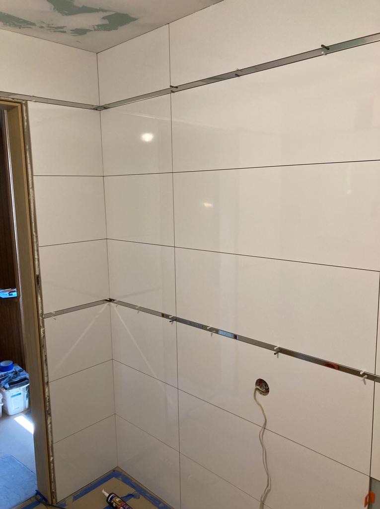 Białe, błyszczące płytki ceramiczne na ścianie łazienki, układane z użyciem metalowych listew poziomujących, widoczne puszki elektryczne i narzędzia glazurnicze na podłodze.