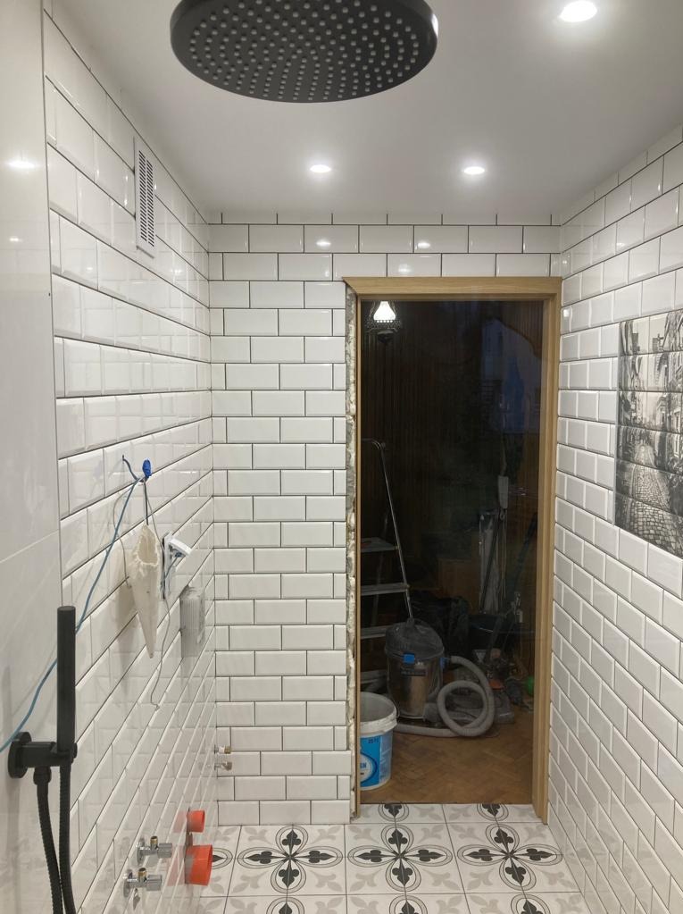 Wnętrze łazienki w trakcie remontu z widocznymi białymi, prostokątnymi płytkami na ścianach, wzorzystą podłogą, czarną deszczownicą oraz otwartym przejściem do innego pomieszczenia, gdzie widoczne...