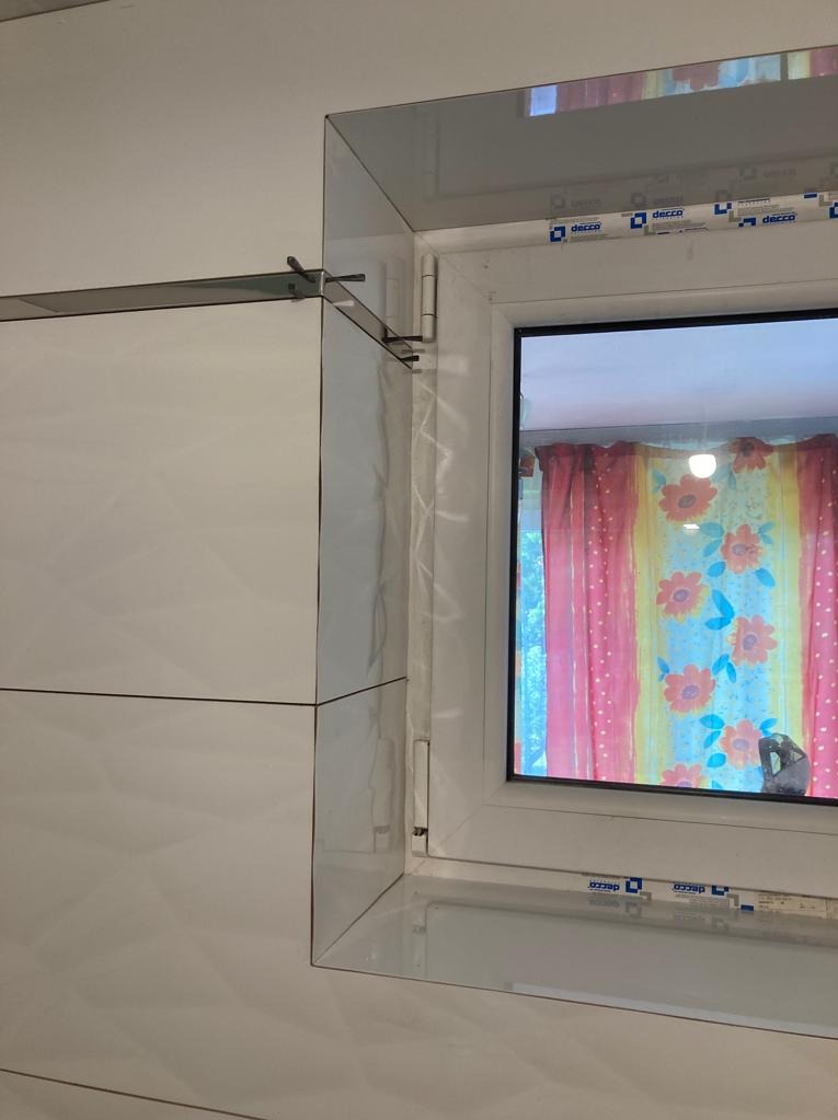 Białe, błyszczące płytki ceramiczne ułożone wokół okna PCV z widocznymi śladami silikonu i uchwytem montażowym, w tle wzorzysta zasłona w odcieniach czerwieni i żółci.