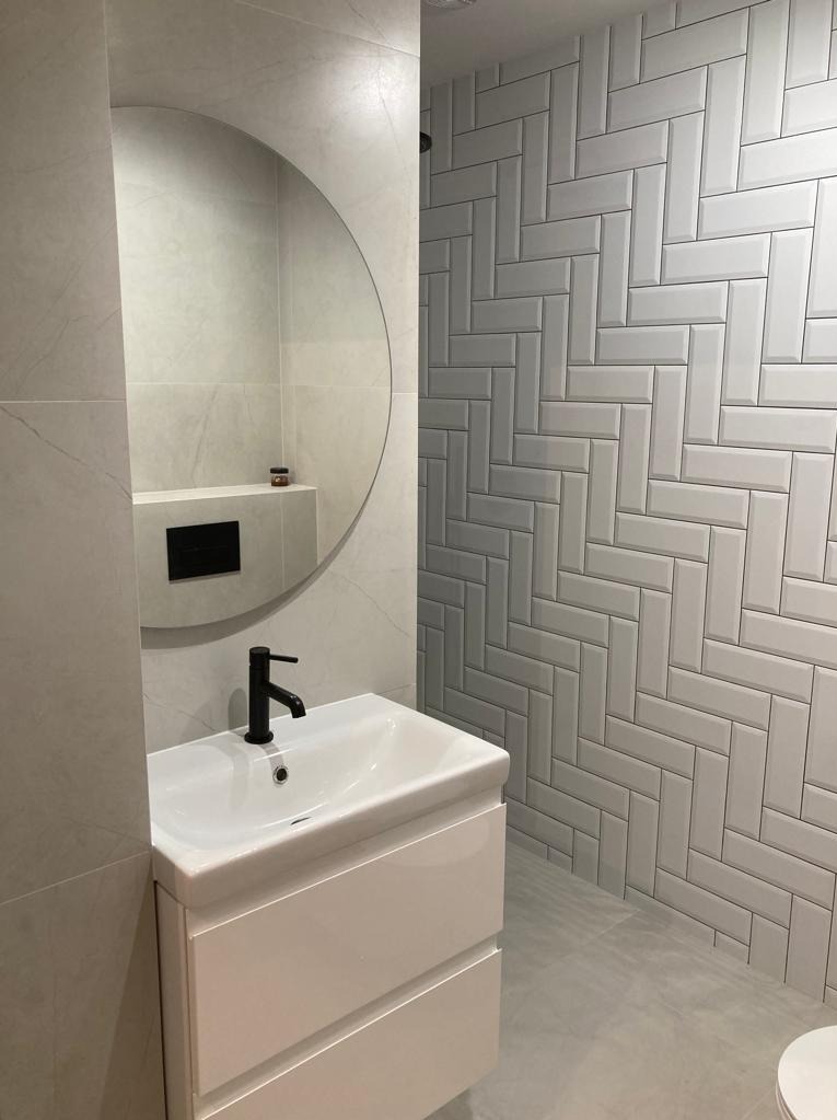 Nowoczesna łazienka z umywalką na szafce, okrągłym lustrem i ścianą wyłożoną białymi, ułożonymi w jodełkę płytkami.