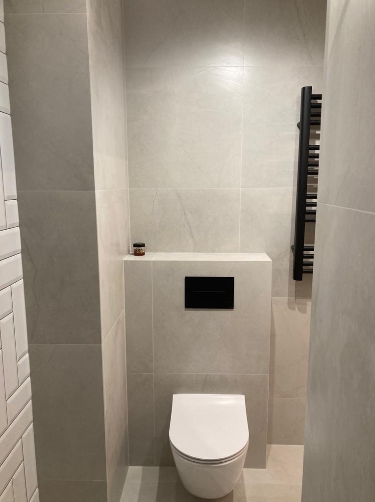 Nowoczesna toaleta podwieszana z czarnym przyciskiem spłukującym, grzejnik drabinkowy w kolorze czarnym oraz płytki imitujące kamień w odcieniach szarości i beżu. Na zabudowie stelaża widoczny mały...