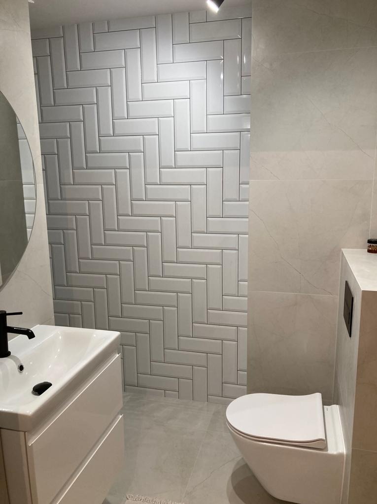 Nowoczesna łazienka z białymi, prostokątnymi płytkami ułożonymi w jodełkę na ścianie, jasnoszara podłoga, biała umywalka z czarną baterią i podwieszana toaleta.