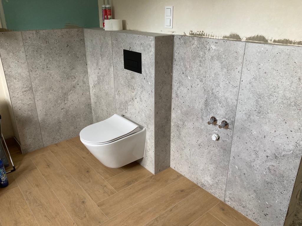 Świeżo wykafelkowana ściana z szarego kamienia w łazience, wisząca toaleta z czarnym przyciskiem spłukiwania, drewnopodobna podłoga ułożona w jodełkę, widoczne przyłącza wodne i ślady po remoncie...