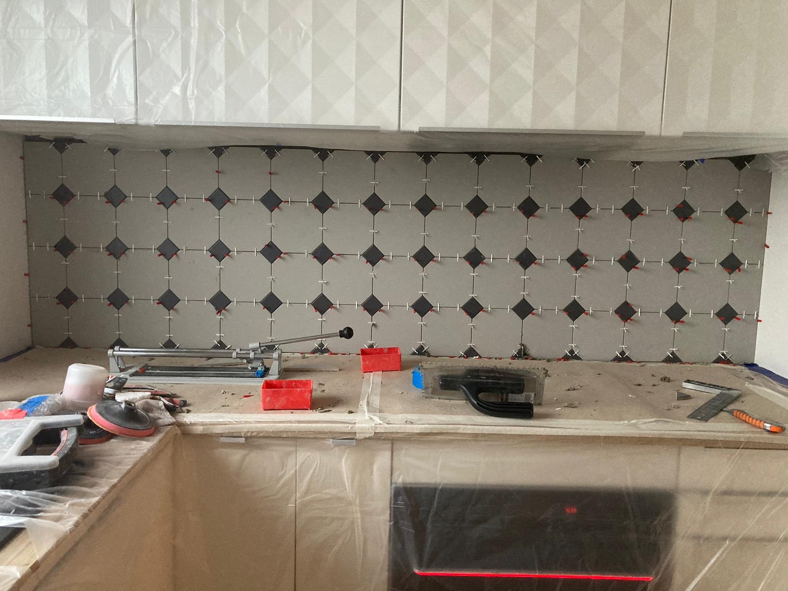 Układanie szaro-czarnych płytek w karo na ścianie w kuchni z użyciem krzyżyków dystansowych, widoczny sprzęt glazurniczy i zabezpieczone folią meble kuchenne.