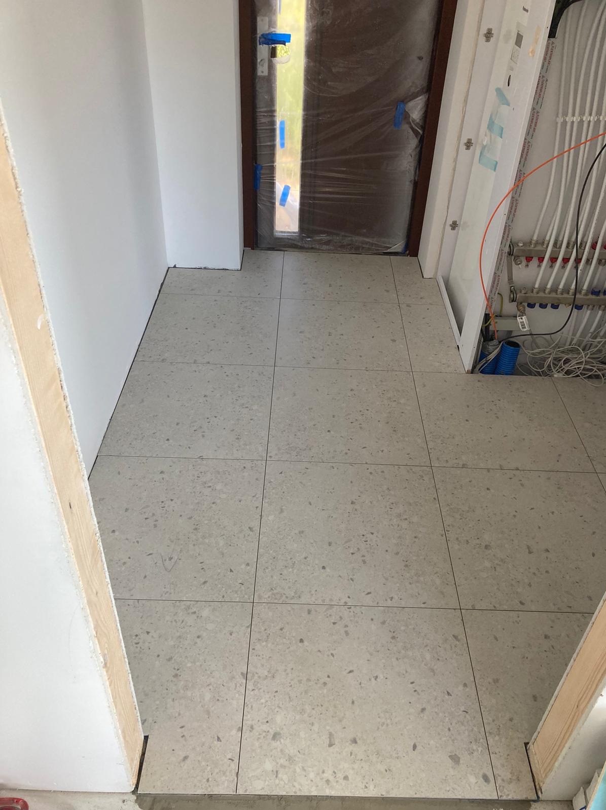 Ułożona jasnoszara terakotowa podłoga w przedpokoju, widoczne fragmenty instalacji grzewczej i zabezpieczone folią drzwi wejściowe.
