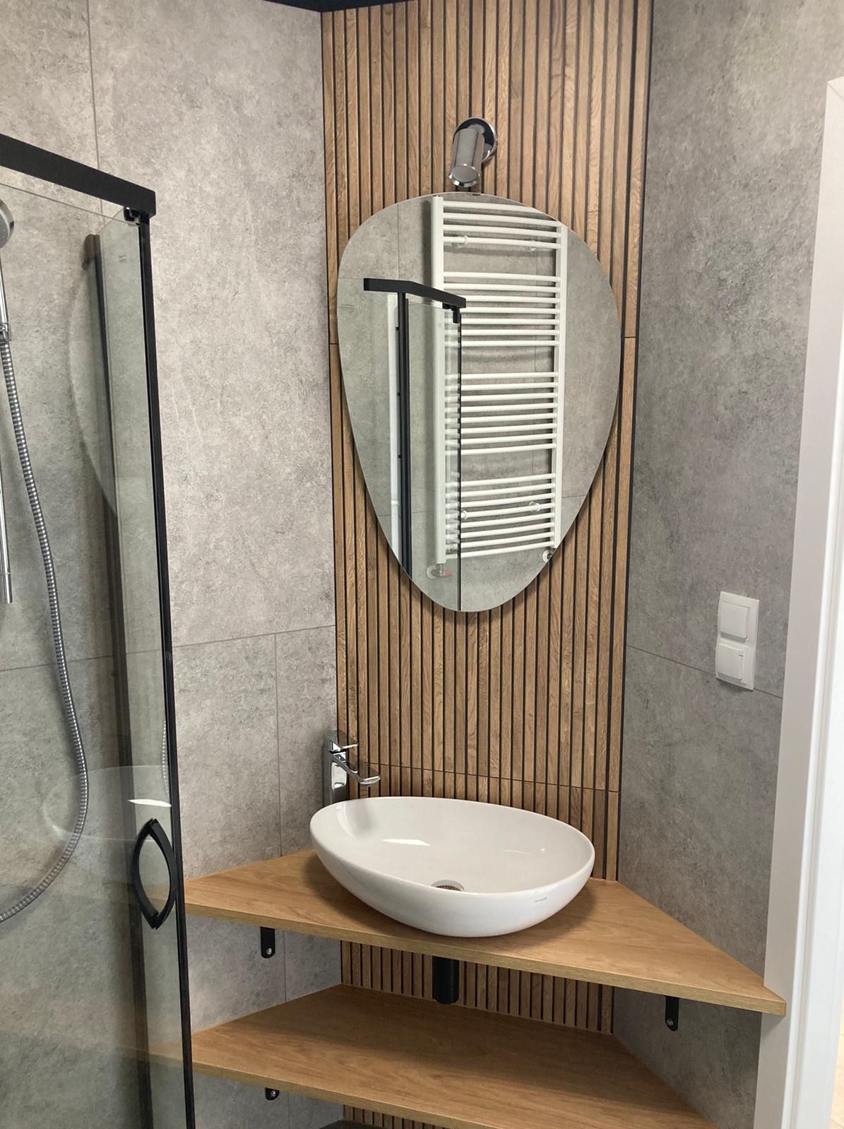 Nowoczesna łazienka z szarą płytką, drewnianymi lamelami i owalnym lustrem, w którym odbija się grzejnik drabinkowy. Umywalka nablatowa na drewnianym blacie narożnym.