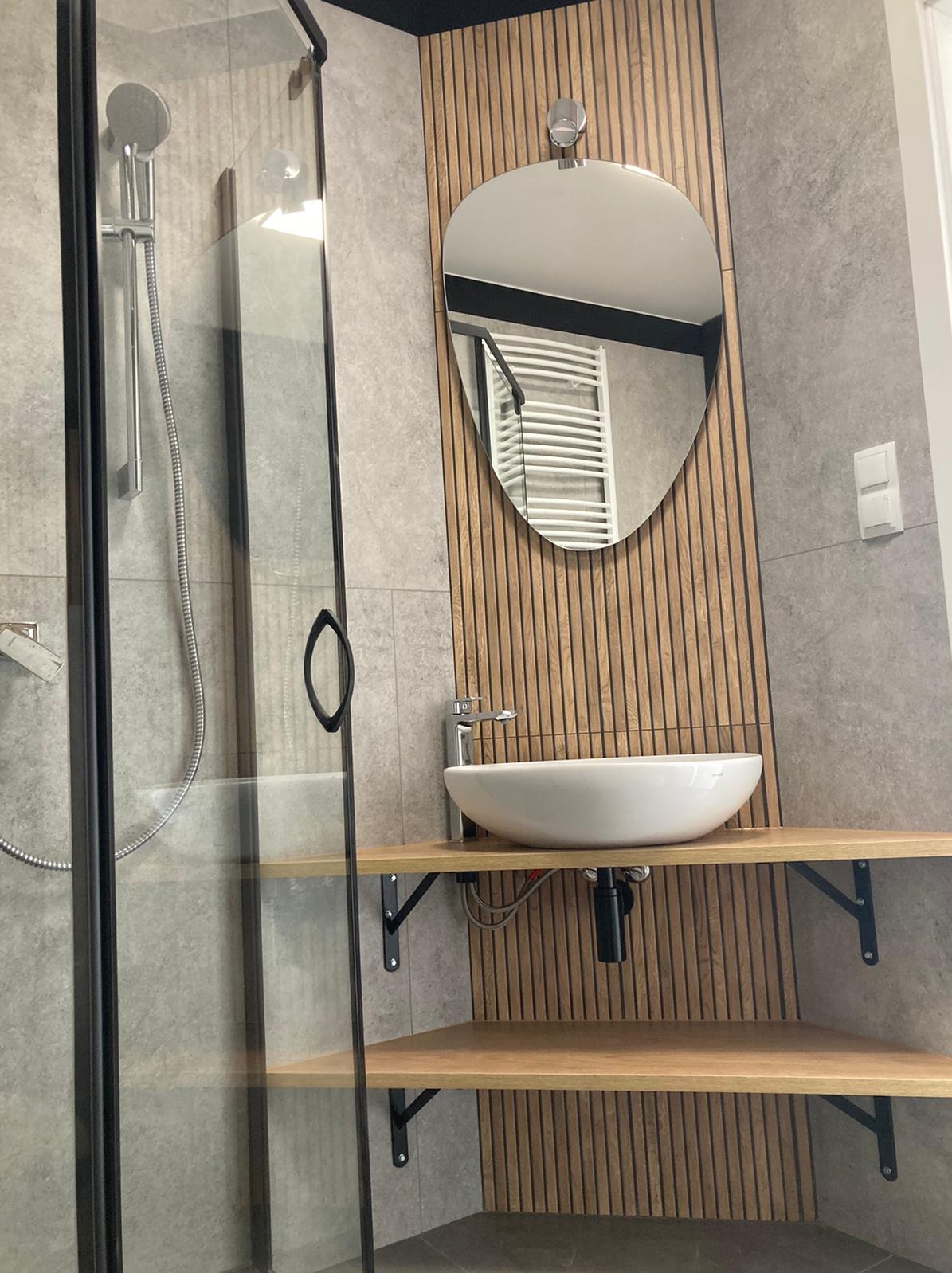 Nowoczesna łazienka z kabiną prysznicową, umywalką nablatową na drewnianym blacie wspartym na czarnych wspornikach, lustrem w kształcie łzy na tle drewnianych paneli ściennych i szarymi płytkami.