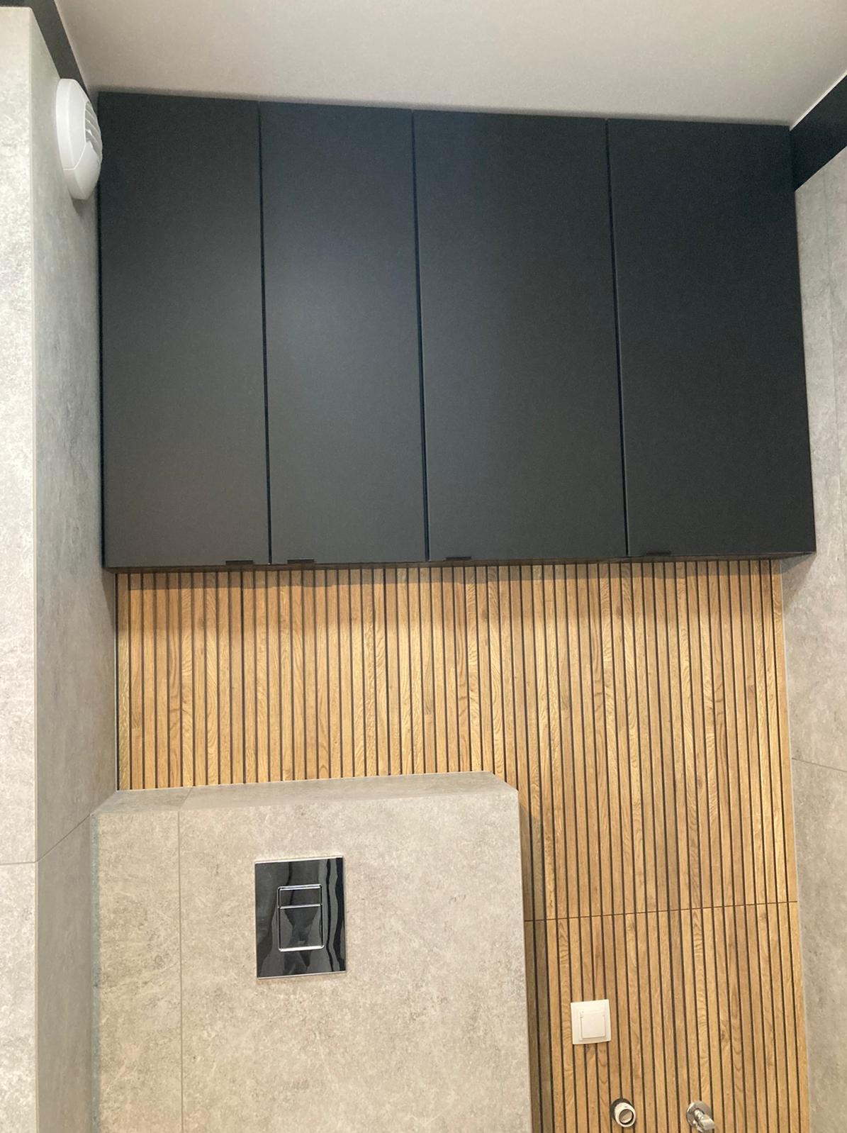 Nowoczesna łazienka po remoncie: szare płytki, drewniana boazeria lamelowa, czarne szafki wiszące i zabudowa toalety podtynkowej z chromowanym przyciskiem.