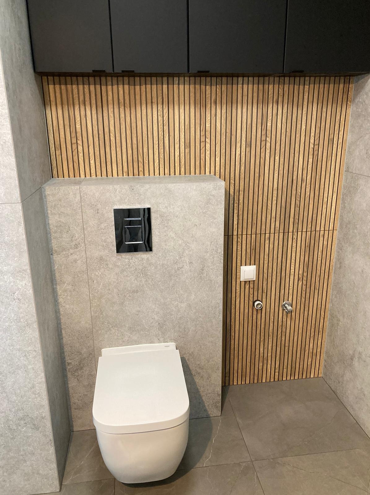 Nowoczesna toaleta podwieszana z szarą płytką i drewnianą ścianką z listew, chromowany przycisk spłukiwania i szare szafki wiszące.