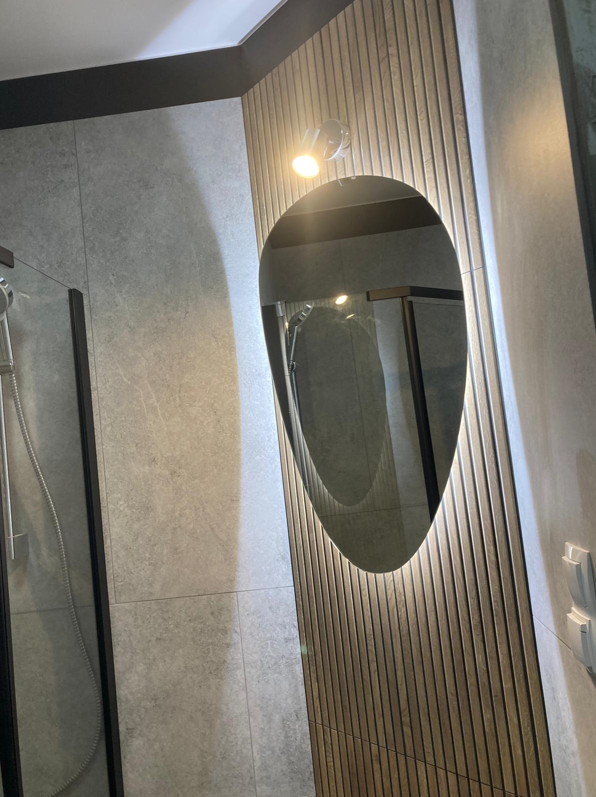 Nowoczesna łazienka z szarą płytką imitującą kamień, kabiną prysznicową z czarnymi profilami i owalnym lustrem z podświetleniem punktowym na ścianie z drewnianych paneli.