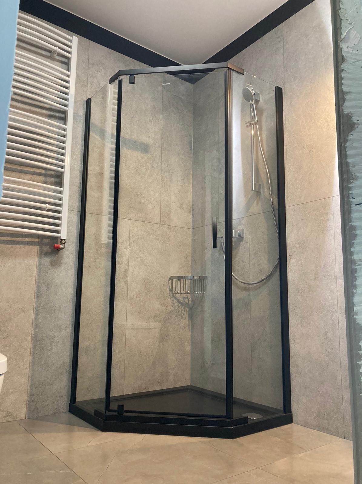 Nowoczesna kabina prysznicowa z czarnymi ramami i szarymi płytkami w trakcie montażu, widoczny fragment nieotynkowanej ściany.