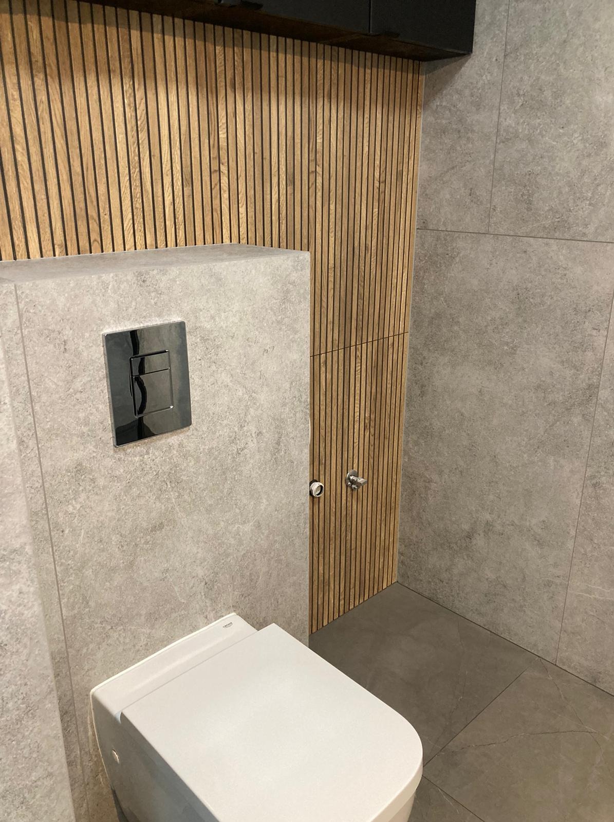 Nowoczesna toaleta podwieszana z chromowanym przyciskiem spłukującym, obudowana płytami imitującymi beton, na tle ściany z drewnianych lameli i płytek w odcieniach szarości.