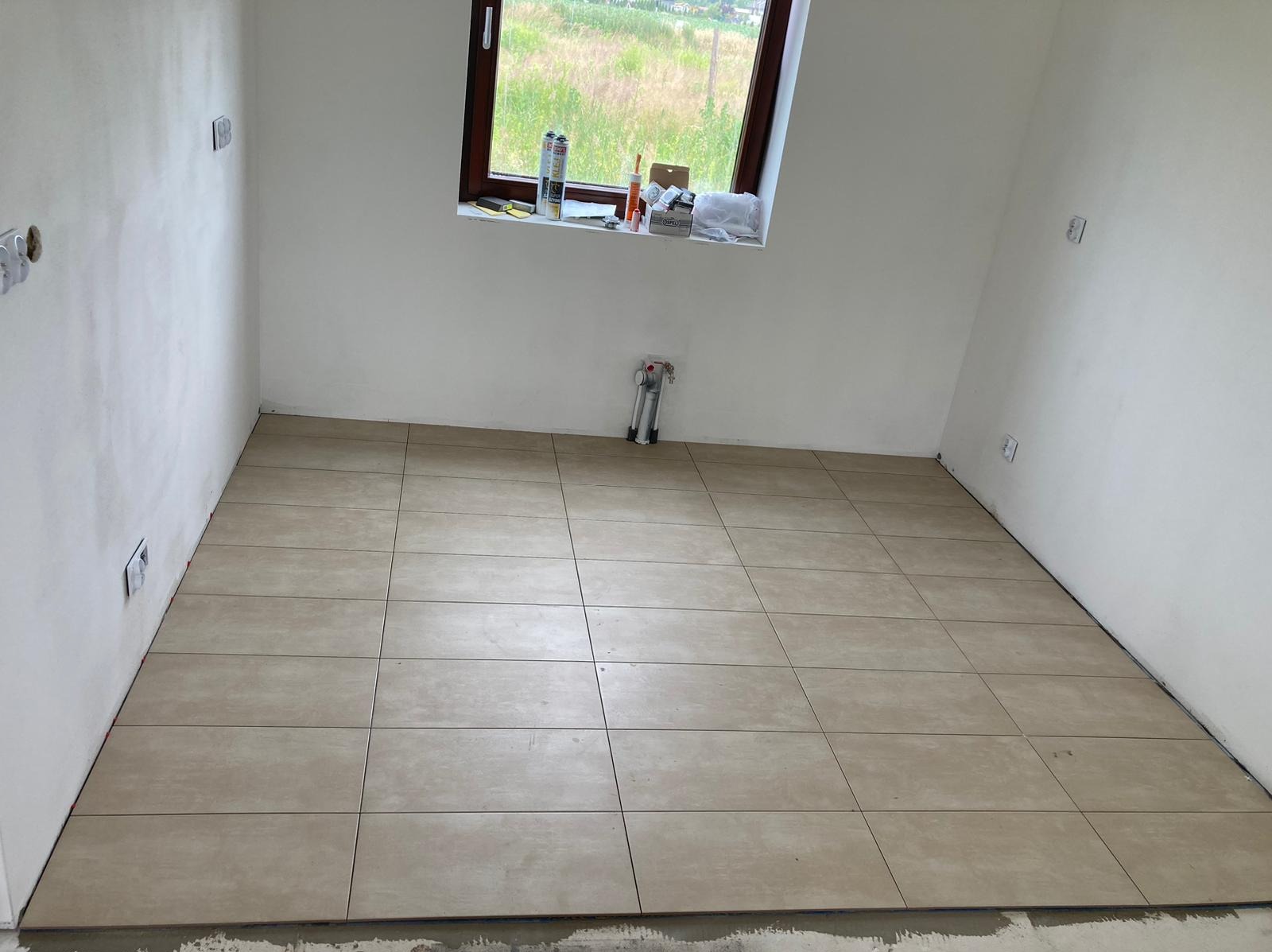 Ułożone beżowe płytki podłogowe w pustym pomieszczeniu z oknem, widoczne puszki z silikonem na parapecie i wystające rury kanalizacyjne ze ściany.