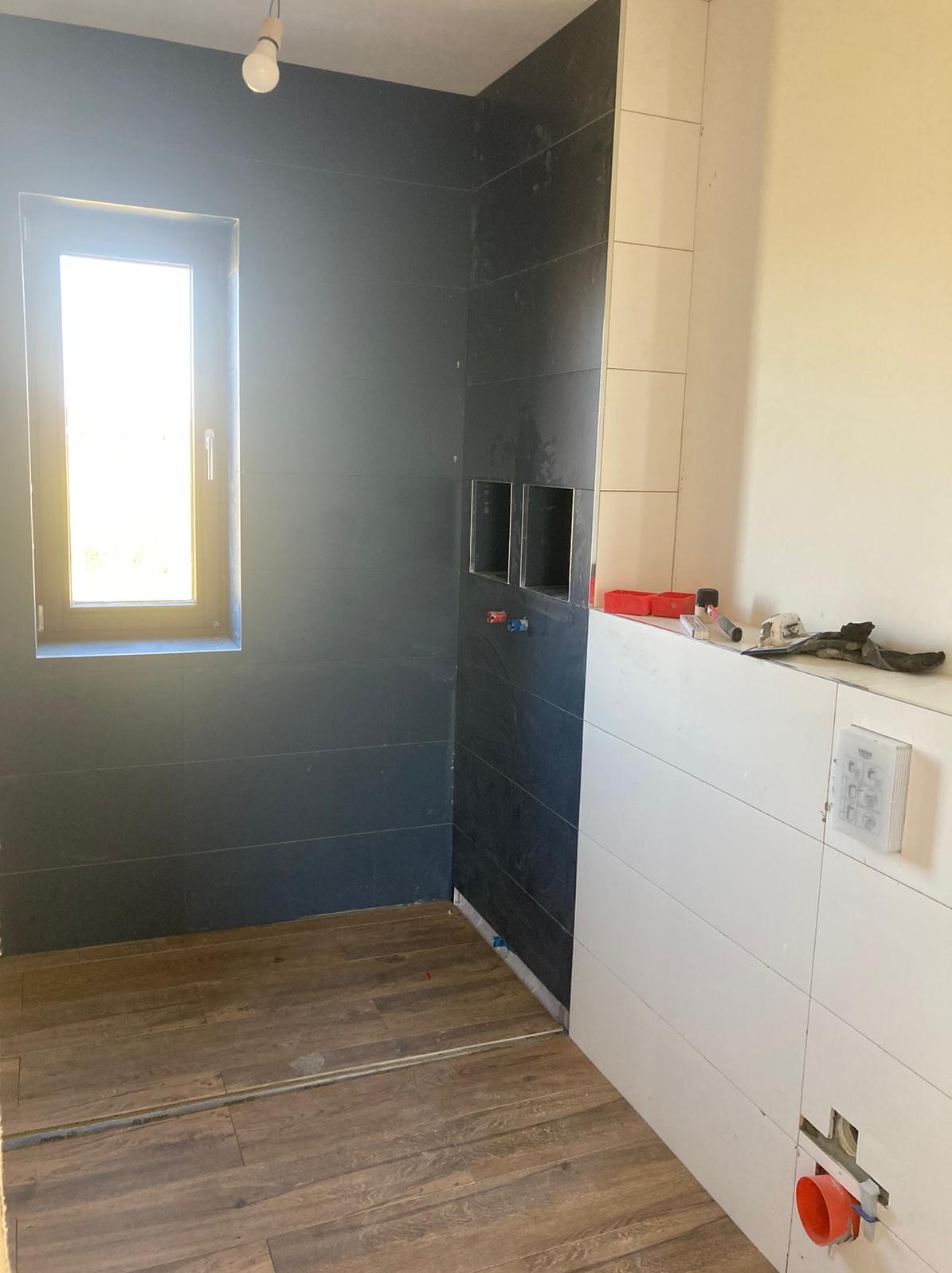 Wnętrze łazienki w trakcie prac wykończeniowych: ułożone płytki w kolorze grafitowym i białym, widoczne przygotowane wnęki, drewnopodobna podłoga, instalacja sanitarna w ścianie, surowa żarówka.