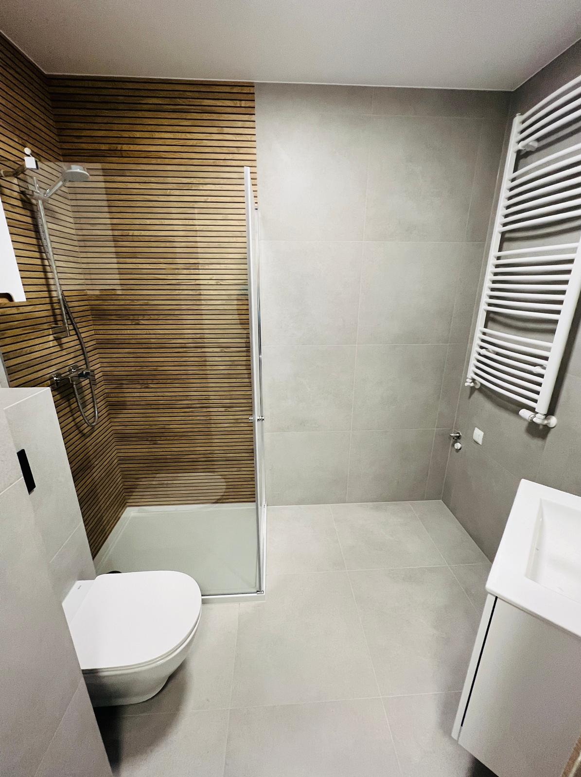 Nowoczesna łazienka po remoncie z kabiną prysznicową, drewnianą okładziną ścienną w strefie prysznica, grzejnikiem drabinkowym i umywalką z szafką w odcieniach szarości.