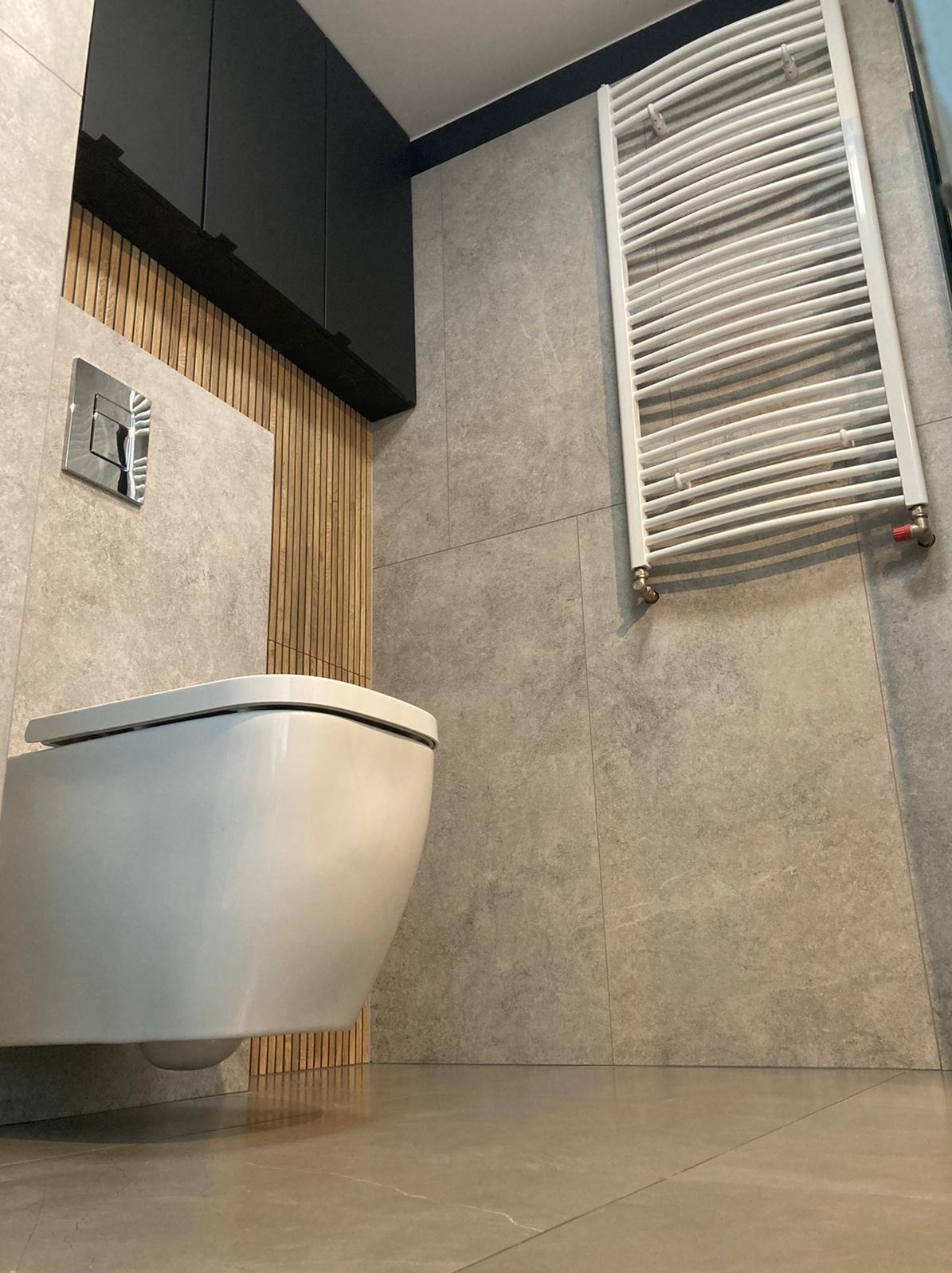 Nowoczesna łazienka z wiszącą toaletą, grzejnikiem drabinkowym i szarą płytką imitującą kamień, oświetlona ciepłym światłem, z elementami drewnianej boazerii.