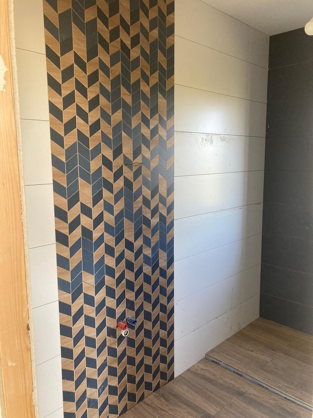 Ściana łazienki w trakcie wykańczania z dekoracyjną mozaiką ułożoną w jodełkę z płytek drewnopodobnych i grafitowych, widoczne przyłącza wodne i fragment podłogi.