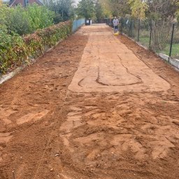 Huzar Maciej Możejko - Przygotowanie podłoża pod ścieżkę z kostki brukowej: ubijanie ziemi zagęszczarką wzdłuż ogrodzenia, wytyczona linia, widoczne ślady opon.