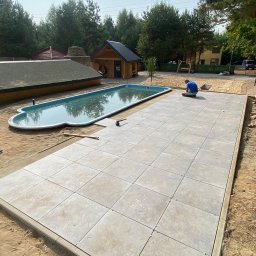 Huzar Maciej Możejko - Układanie jasnoszarej kostki brukowej na piasku obok basenu w ogrodzie, widoczne krzyżyki dystansowe, narzędzia i pracownik w trakcie pracy.